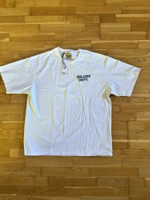 Vit Gallery Dept. t-shirt - Snygg vit t-shirt från Gallery Dept. med svart tryck fram och stort tryck på ryggen där det står 'GALLERY DEPT. HOLLYWOOD, CA.'. Klassisk passform och rund hals. Perfekt för dig som gillar streetwear och enkel stil.