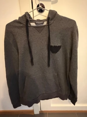 Grå hoodie från Emporio Armani - Mysig grå hoodie från Emporio Armani med svart logga på bröstet och klassisk känguruficka. Hoodien har dragsko i huvan och ribbade muddar. Perfekt för en chill och stilren look. Materialet är mjukt och fleeceliknande på insidan.