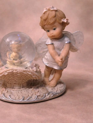 Dekorativ ängel med snöglob - Söt prydnadsfigur föreställande en ängel i vit klänning med glitterdetaljer och vingar, bredvid en snöglob med en liten figur inuti. Basen är dekorerad med blommor och mönster i ljusa toner. Perfekt som dekoration i hemmet.