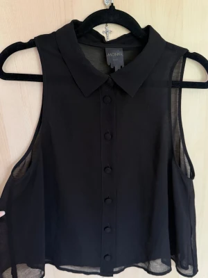 Svart blus från Monki - Stilren svart ärmlös blus från Monki med klassisk krage och knappar framtill. Tunt och lätt material som ger en luftig känsla, perfekt för varmare dagar. Enkel att matcha med olika outfits och passar dig som gillar minimalistisk stil.