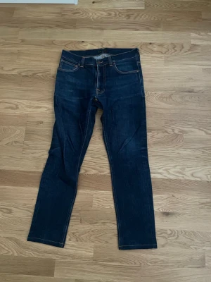 Mörkblå skinny jeans från Nudie - Snygga mörkblå skinny jeans från Nudie med klassiska orangea sömmar och broderade bakfickor. Jeansen har fem fickor, låg midja och är tillverkade i stretchigt denim för en bekväm passform. Perfekta för en trendig och avslappnad look.