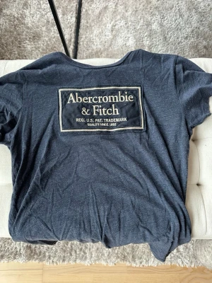 Mörkblå Abercrombie & Fitch t-shirt - Snygg mörkblå t-shirt från Abercrombie & Fitch med stort logotryck på ryggen. Klassisk passform och mjukt material som känns skönt mot huden. Perfekt för dig som gillar stilrena och tidlösa plagg.