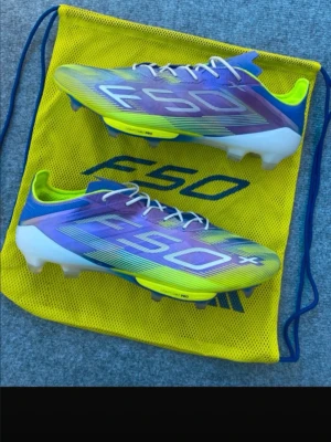 Adidas F50 fotbollsskor lila/gul - Säljer ett par Adidas F50 fotbollsskor i en snygg mix av lila, blått och neon-gult med vita detaljer. Skorna har låg profil, snörning och är tillverkade i lätt syntetmaterial för maximal fart på planen. Perfekta för dig som vill sticka ut och spela snabbt.