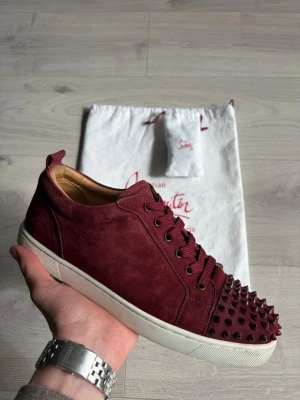 Louis Junior Suade - Vinröda sneakers från Christian Louboutin med nitar. Skorna är i generellt mycket fint skick och det enda tecknet på användning är smuts under sulan. Skorna säljs med en dustbag och extra original skosnören.