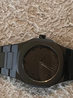 Svart stilren klocka i metall - Snygg svart klocka med minimalistisk design och mattsvart urtavla. Klockan har metallarmband och stilrena detaljer utan siffror, vilket ger ett modernt och clean intryck. Perfekt accessoar för dig som gillar en diskret men cool look.