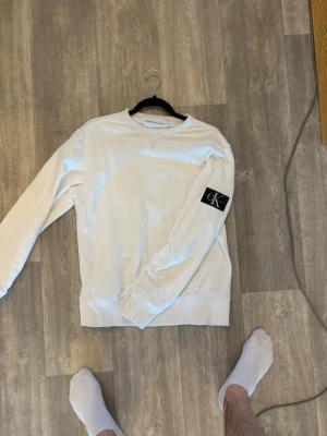 Vit sweatshirt från Calvin Klein - Vit sweatshirt från Calvin Klein med rund halsringning och ribbade muddar. Tröjan har en svart patch med CK-logga på vänster ärm och är tillverkad i mjukt bomullsmaterial. Enkel och stilren design som passar perfekt till jeans eller joggers. Storlek S