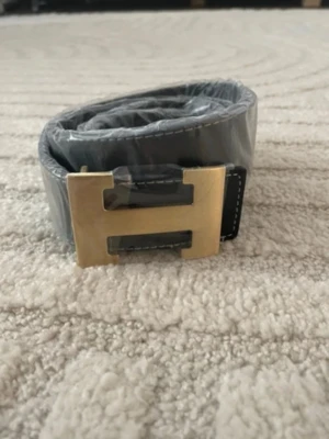 Svart läderbälte med H-spänne - Snyggt svart bälte i läder med ett stort, guldigt H-format metallspänne. Bältet har vita sömmar längs kanterna som ger en schysst kontrast. Perfekt accessoar för att lyfta din outfit och ge en exklusiv känsla.