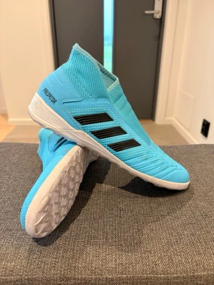 Adidas Predator blå fotbollsskor - Snygga Adidas Predator fotbollsskor i ljusblått med svarta ränder på sidan och vit sula. Skorna har en strumpliknande passform utan snörning och är tillverkade i syntetmaterial, perfekta för inomhusspel. Finns små märken av användning men inget som är sönder eller förstör användningen. OBS!! Skorna finns även ute på Vinted. 