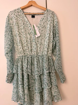 Grön blommig volangklänning Ginatricot - Supersöt kort klänning från Vero Moda i ljusgrönt med småblommigt mönster. Klänningen har v-ringning, långa ärmar och flera volanger i kjolen för en flowy look. Perfekt för vår och sommar!