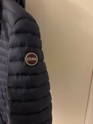 Mörkblå pufferjacka från Colmar - Snygg mörkblå pufferjacka från Colmar med huva och dragkedja framtill. Jackan har två fickor med dragkedja och ett diskret Colmar-märke på ärmen. Perfekt för kalla dagar tack vare det vadderade materialet och den stilrena designen. Den ser lite skrynklig ut men de är för den legat ner packad