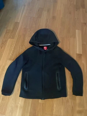 Svart hoodie från Nike med dragkedja - Snygg svart hoodie från Nike med dragkedja framtill och två fickor med dragkedja. Klassisk huva och diskret design utan tryck. Tillverkad i mjukt material som känns bekvämt mot huden. Perfekt för dig som gillar sportig och enkel stil.