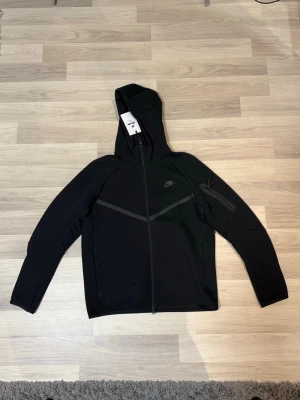 Svart Nike tech fleece  - Snygg svart Nike tech fleece hoodie med hel dragkedja framtill och huva. Diskret Nike-logga på bröstet och en cool detaljficka på ena ärmen. Helt ny, köpt i Paris som present men eftersom att den aldrig använts så säljer jag nu vidare för billigare 