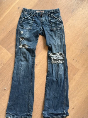 Blå slitna jeans med vida ben - Säljer ett par blå jeans med vida ben och flera slitningar på framsidan. Jeansen har stora fickor både fram och bak samt detaljerade sömmar. Perfekta för en avslappnad och trendig streetstil. Priset kan diskuteras