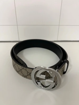 Gucci GG Supreme bälte med silverdetalj - Snyggt Gucci GG Supreme bälte med klassiskt monogrammönster i beige och brunt. Bältet har en stor, rund spänne i silverfärgad metall med dubbla G-loggan. Tillverkat i läder och canvas, perfekt för att lyfta vilken outfit som helst.