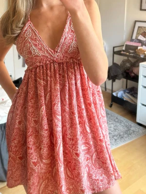 Rosa/röd kort klänning - Supersöt kort klänning i rosa och vitt med paisleymönster och v-ringad omlott upptill. Klänningen har tunna axelband och ett luftigt, flowy fall. Perfekt för varma dagar och ger en bohemisk vibe.