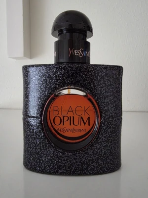 Yves Saint Laurent Black Opium parfym - OBS Kolla min annons med samma parfym och gratis frakt 💟💟💟Black Opium från Yves Saint Laurent är en ikonisk parfym i en svart, glittrig flaska med rund orange detalj och svart kork. Flaskan har en modern, lyxig design och är tillverkad i glas med en glittrig finish. Perfekt för dig som gillar djärva och eleganta dofter. 