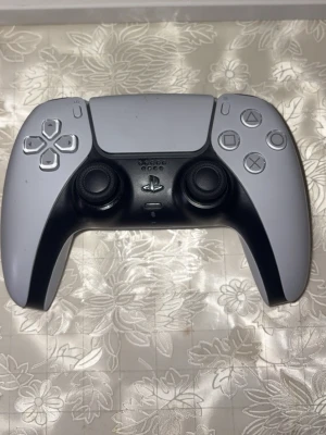 PlayStation 5 DualSense handkontroll - Säljer en PlayStation 5 DualSense handkontroll från Sony. Framsidan är vit med svarta detaljer och har klassiska PlayStation-knappar. Baksidan har ett grått kamouflagemönster. Ergonomisk form och tillverkad i plast, perfekt för gaming.