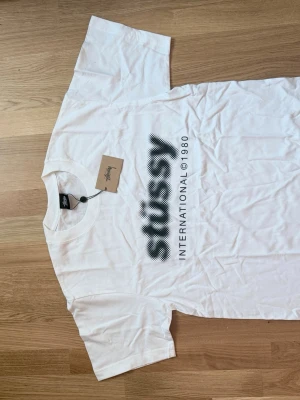Vit Stussy t-shirt med tryck - Vit t-shirt från Stussy med svart grafiskt tryck på framsidan där det står 'Stussy International © 1980'. Klassisk passform och rund halsringning. Perfekt för en streetwear-look.