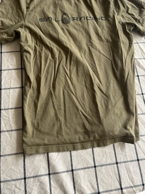 Olivgrön t-shirt från Sail Racing - Olivgrön t-shirt från Sail Racing med svart logga tryckt på ryggen. Klassisk passform och korta ärmar, perfekt för en avslappnad stil. Tillverkad i mjuk bomull som känns skön mot huden.