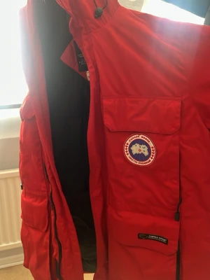 Röd vindjacka från Canada Goose - Riktigt snygg röd vindjacka från Canada Goose med huva och flera stora fickor framtill. Jackan har klassisk logga på bröstet och svarta muddar vid ärmsluten. Perfekt för kalla och blåsiga dagar, med en stilren och sportig look.