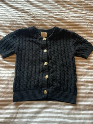 Svart kabelstickad kofta med kort ärm - Svart kabel stickad kofta i storlek Xs från H&M. Perfekt till både sommar och vinter, perfekt till sommarkvällar när det är lite kyligt men ändå varmt. Den har snygga guldknappar, supersnyggt att matcha med guldsmycken, eller silversmycken.