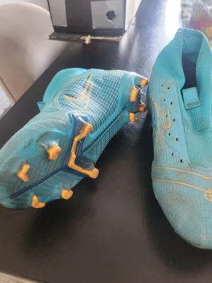 Nike blå fotbollsskor med dobbar - Säljer ett par blå Nike fotbollsskor med gula detaljer och dobbar. Skorna har en strömlinjeformad design utan synliga snören och är tillverkade i syntetmaterial för bra grepp och kontroll på planen. Perfekta för dig som vill sticka ut på fotbollsplanen.