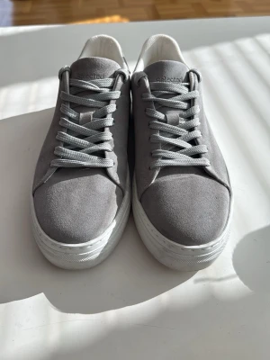 Gråa sneakers från Selected Homme - Stilrena gråa sneakers från Selected Homme med ovandel i mockaliknande material och vita tjocka sulor. Klassisk låg modell med matchande grå skosnören och diskret logga på plösen. Perfekta för en clean och modern look. Helt nya använda 1 gång.