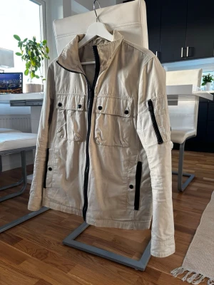 Beige vårjacka med svarta detaljer - Stilren beige vårjacka med svarta dragkedjor och knappar. Jackan har hög krage, fyra fickor framtill och en extra ficka på ärmen. Lätt material och rak passform, perfekt för blåsiga dagar. Några få defekter som visas på bild 4 och 5. Hör av er vid frågor!