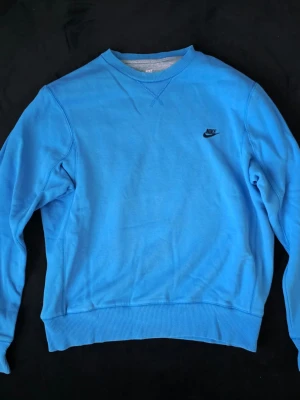 Blå sweatshirt från Nike - Snygg blå crewneck från Nike med svart broderad logga på bröstet. Tröjan har rund halsringning, ribbade muddar och är tillverkad i mjukt material. Perfekt för en avslappnad och sportig stil.