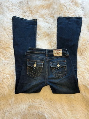 True Religion mörkblå bootcut jeans - Mörkblå bootcut jeans från True Religion i design Becca med snygga kontrastsömmar och dekorativa ficklock bak. Klassisk femficksmodell med knappar och dragkedja framtill. Ikoniska detaljer på bakfickorna och patch med logga i midjan. Perfekta för en trendig och avslappnad look. Lite slitna längst ner men ingen som syns då de används❣️
