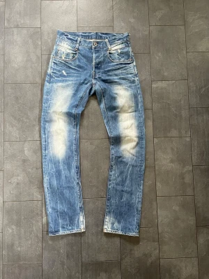 Blå slitna jeans med ljusa partier - Säljer ett par blå jeans med coola slitningar och ljusa partier på lår och knän. Jeansen har klassisk femficksdesign, raka ben och tydliga kontrastsömmar. Perfekta för en avslappnad streetstil.