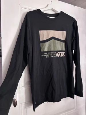 Svart långärmad t-shirt från Vans - Svart långärmad tröja från Vans med grafiskt tryck i beige och grönt på bröstet. Klassisk passform och rund halsringning. Trycket har texten 'The Original Established 1966 Off The Wall'. Perfekt för en avslappnad streetstil.