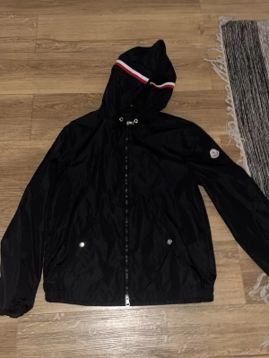 Svart Moncler vindjacka med huva - Svart vindjacka från Moncler med huva och dragkedja framtill. Jackan har två fickor med tryckknappar och Moncler-logga på ärmen. Huvan har en snygg detalj med röd och vit rand. Perfekt för blåsiga dagar och streetwear-stil.