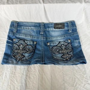 Blå jeanskjol med broderade fickor - Snygg blå jeanskjol med kort modell och två bakfickor dekorerade med broderade mönster och nitar. Kjolen har bälteshällor och slitna detaljer för en cool look. Perfekt för dig som gillar streetstyle.