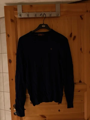 Mörkblå tröja med rund hals Morris  - Mörkblå långärmad tröja med rund hals och diskret broderad logga på bröstet. Tröjan har ribbade muddar vid ärmslut och nederkant. Perfekt för en stilren och enkel look.