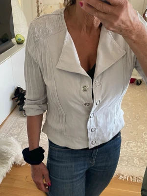 KRISS ljusgrå jacka, storlek S - KRISS ljusgrå jacka, storlek S Ljusgrå kofta med snygga knappar framtill och unik struktur på axlar och rygg. Modellen har 3/4-ärmar och en bred krage som ger en cool look. Materialet är mjukt och bekvämt, perfekt för lager-på-lager-stil.