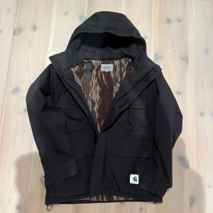 Svart jacka från Carhartt WIP - Säljer en svart jacka från Carhartt med huva och dragkedja. Jackan har två stora fickor framtill och ett camouflagemönstrat foder. Perfekt för höst och vinter nypris ca 2500kr. Helt ny tvättad från kemtvätt  och inte använd efter det. Kan säljas billigare vid snabb affär. Har tagit bort fickorna vid bröstet så kan tänka mig att sälja den billigare för de är små hål men det går att sy 