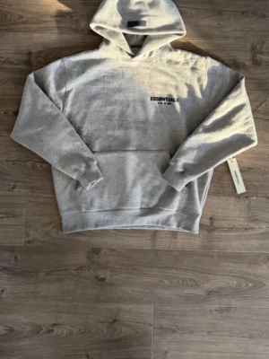 Grå Essentials Fear of God hoodie - Helt ny Grå Essentials Fear of God hoodie. Aldrig använd och kommer med lapp. Säljer den eftersom den är för stor för mig. Den är S men lite baggy och passar som typ M. Pris kan diskuteras vid snabba affärer.