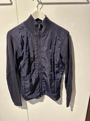 Mörkblå cardiganjacka från Gant - Mörkblå cardiganjacka från Gant med stickade ärmar och rygg samt glansig framsida i nylon. Jackan har hög krage, dragkedja framtill och diskret logga på bröstet. Perfekt för lager på lager och en clean look.