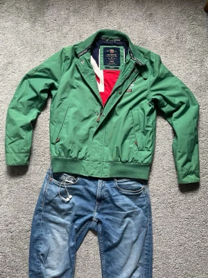 Grön vindjacka från Morris - Snygg grön vindjacka från Morris med dragkedja, ribbade muddar och broderade detaljer på bröstet. Jackan har två fickor med knappstängning och en klassisk krage. Perfekt för vår och höst, lätt att matcha med jeans.