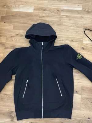Svart vindjacka från Stone Island - Svart vindjacka med huva från Stone Island. Jackan har två framfickor med dragkedja, hel dragkedja framtill och den klassiska Stone Island-loggan på vänster ärm. Materialet är tunt och smidigt, perfekt för blåsiga dagar. Köpt på NK för 7 tusen. Jackan är i bra skick 