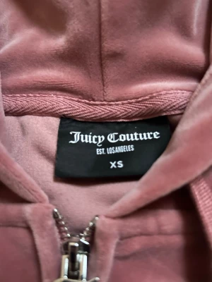 Rosa velourhoodie från Juicy Couture - Mysig rosa hoodie i velour från Juicy Couture med dragkedja framtill, huva och kängurufickor. Klassisk logga broderad på bröstet och ribbade muddar vid ärmslut och nederkant. Perfekt för en chill och trendig look.