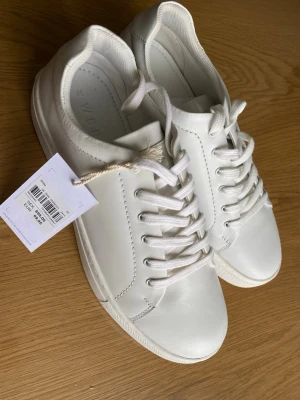 Vita sneakers från Weekday - Stilrena vita sneakers från Weekday med klassisk låg profil och vita snören. Skorna har en slät ovandel i syntetmaterial och vit sula. Perfekta för en clean och enkel look.
