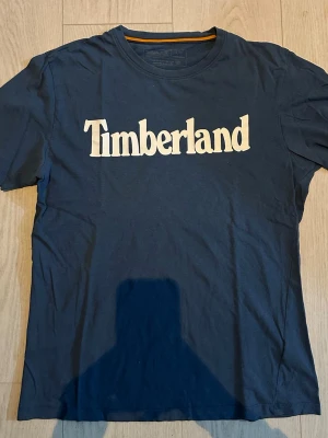 Mörkblå Timberland t-shirt - Snygg mörkblå t-shirt från Timberland med stor vit logga framtill. Klassisk passform och rund halsringning. Perfekt för en avslappnad och stilren look.