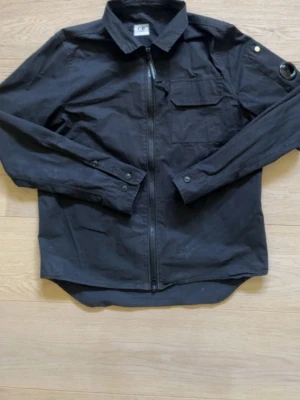 Svart overshirt från C.P. Company - Stilren svart overshirt från C.P. Company med dragkedja framtill, klassisk krage och bröstficka. Långärmad modell med knappdetaljer vid ärmslut och ikonisk lins på ena ärmen. Perfekt lager-på-lager-plagg i bomull.