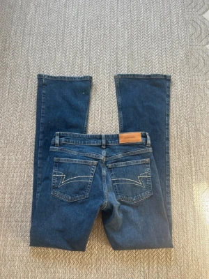 Mörkblå bootcut jeans från Junkyard - Snygga mörkblå bootcut jeans från Junkyard med klassiska fem fickor och kontrastsömmar på bakfickorna. Jeansen har en låg midja och är tillverkade i denim av bomull. Perfekta för en avslappnad och trendig look.