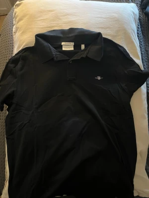 Svart pikétröja från GANT XL - Klassisk svart pikétröja från GANT i regular fit. Tröjan har krage, knappar framtill och GANT:s broderade logga på bröstet. Perfekt till jeans eller chinos för en clean och stilren look.