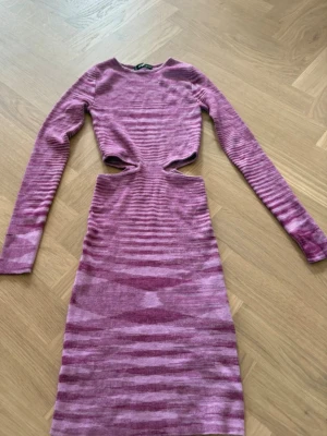 Långärmad rosa stickad klänning med cut-outs - Säljer en långärmad stickad klänning i rosa toner med coola cut-outs vid midjan. Klänningen har ett randigt mönster och är figurnära med en längre passform. Perfekt för dig som vill sticka ut med en unik look.