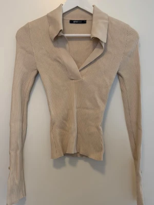 Beige ribbad tröja med krage - Trendig beige ribbad tröja från Gina Tricot med djup v-ringning och krage, öppen nedtill i ärmar med en liten knapp. Långärmad modell i stretchigt material som sitter åt och formar  kroppen.  Inga fler bilder kan slickas då den ligger i flyttlåda.