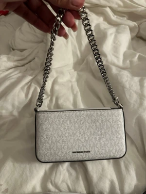 Vit handväska Michael Kors  - Snygg vit handväska från Michael Kors med silverfärgad kedjerem och avtagbar axelrem i läder. Väskan har en stilren, rektangulär form och diskret logga framtill. Materialet är präglat konstläder som ger en lyxig känsla. Perfekt accessoar för att lyfta din outfit. Helt ny, kommer tyvärr inte till användning. Köpt för 1400kr 